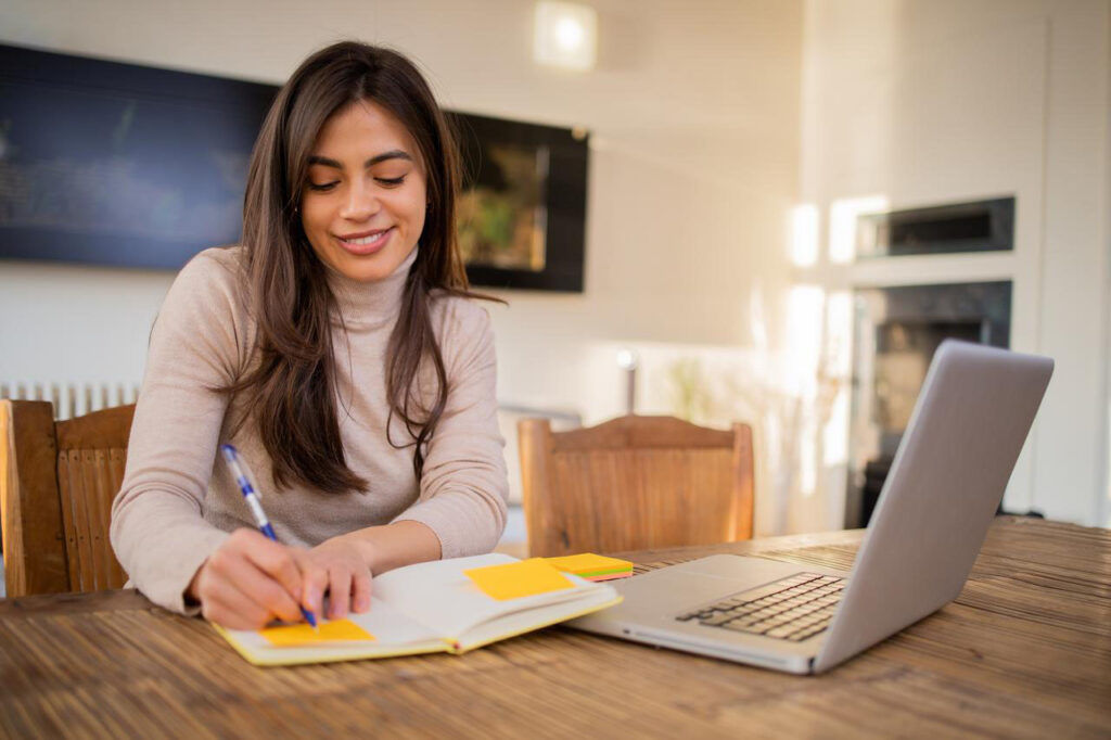 A woman studying at a wooden table with a laptop and notebook reviews example real estate exam questions for her Ohio real estate exam.